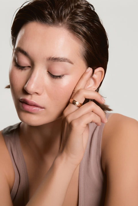 Woman with eyes closed wearing a gold stacked ring near her cheek, head slightly bowed, in a mauve tank top