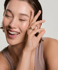 Smiling woman wearing a gold band ring on her finger, touching her cheek, wearing a mauve tank top against a neutral background