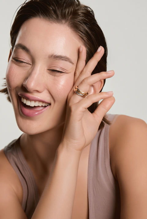 Smiling woman wearing a gold band ring on her finger, touching her cheek, wearing a mauve tank top against a neutral background
