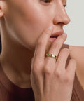 Close-up of a person wearing a gold ring with green gemstones, touching their face.