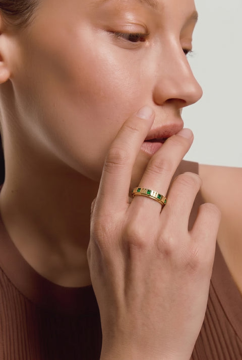 Close-up of a person wearing a gold ring with green gemstones, touching their face.