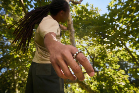 Person with dreadlocks holding a small object outdoors with trees in the background
