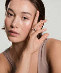 A woman wearing a beige tank top is holding her hand to the side of her head, displaying a rose gold spinner anxiety ring on her finger encrusted in crystals. 