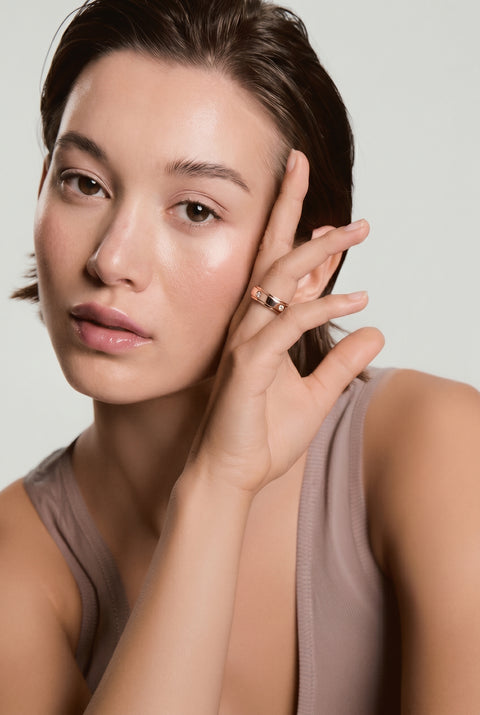 A woman wearing a beige tank top is holding her hand to the side of her head, displaying a rose gold spinner anxiety ring on her finger encrusted in crystals. 