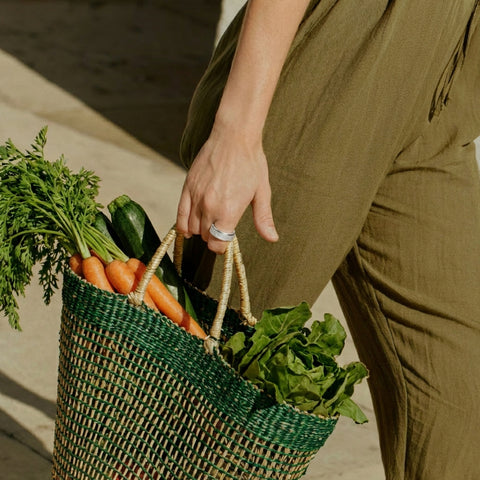 Person holding a green woven bag filled with fresh vegetables.
