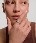 Woman with short hair looking upward, wearing a silver diamond-encrusted spinning ring on her finger, touching her cheek in a brown ribbed tank top