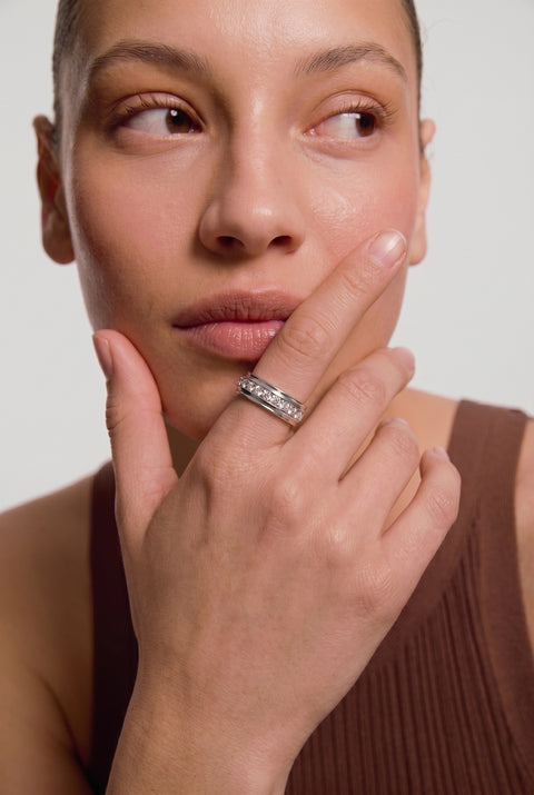 Woman with short hair looking upward, wearing a silver diamond-encrusted spinning ring on her finger, touching her cheek in a brown ribbed tank top