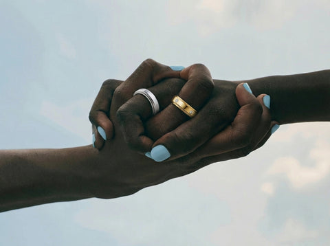 Two hands holding each other, one with light blue nail polish and a silver ring and one with a gold ring, against a sky background